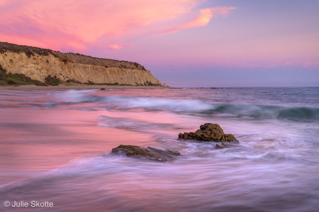 Sunset at Crystal Cove State Park in California. The sky and water is colored pink and orange flow the setting sun. The high hide tide and waves covers the beach.