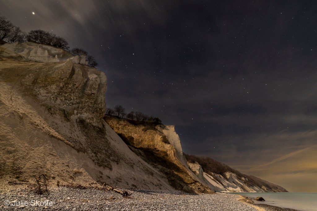 Måneskinnet oplyser Møns klint, mens en mørk nattehimmel med stjerner ses i baggrunden.