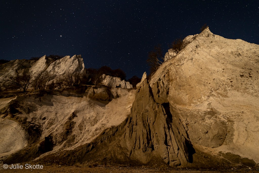 Måneskinnet oplyser Møns klint, mens en mørk nattehimmel med stjerner ses i baggrunden.