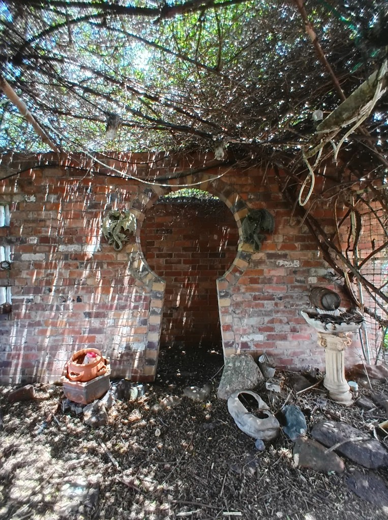 This week’s photo shows a brick enclosure with an opening in the wall that looks like a large keyhole leading to another brick enclosure. Streaks of light seem to be coming through the perhaps grass ceiling. There are large stones on the ground and a bird bath in the right corner. It also looks like there are other little treasures on the ground.
