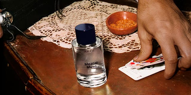 Bottle labeled "Mezcal Musc" next to a hand holding playing cards. Bottle labeled "Mezcal Musc" next to a hand holding playing cards.