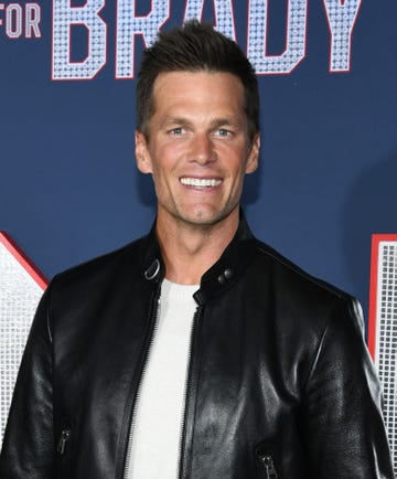 Tom Brady in January 2023 tom brady smiles at the camera, he wears a black leather jacket over a white shirt and stands in front of a navy background