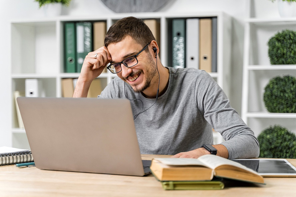 Photograph of a man wearing glasses and headphones smiling while looking at a laptop screen. He is sitting at a table with a book, notebooks and a pen. In the background, there are shelves with folders and potted plants. Photograph of a man wearing glasses and headphones smiling while looking at a laptop screen. He is sitting at a table with a book, notebooks and a pen. In the background, there are shelves with folders and potted plants.