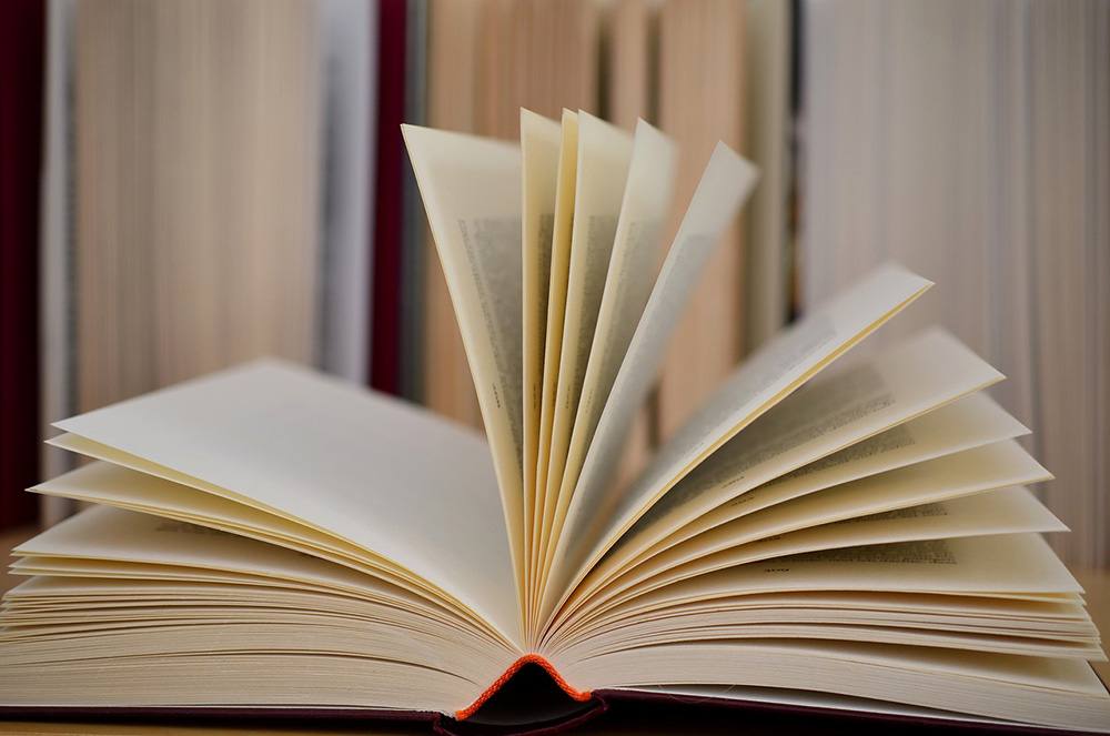 Photo of an open book on a table, in the background there's a bookcase full of books Photo of an open book on a table, in the background there's a bookcase full of books