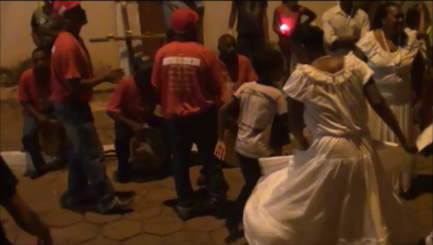 Photograph of a group of Black people participating in a Suça circle. The men are wearing red t-shirts, while the women are in long white dresses. Photograph of a group of Black people participating in a Suça circle. The men are wearing red t-shirts, while the women are in long white dresses.