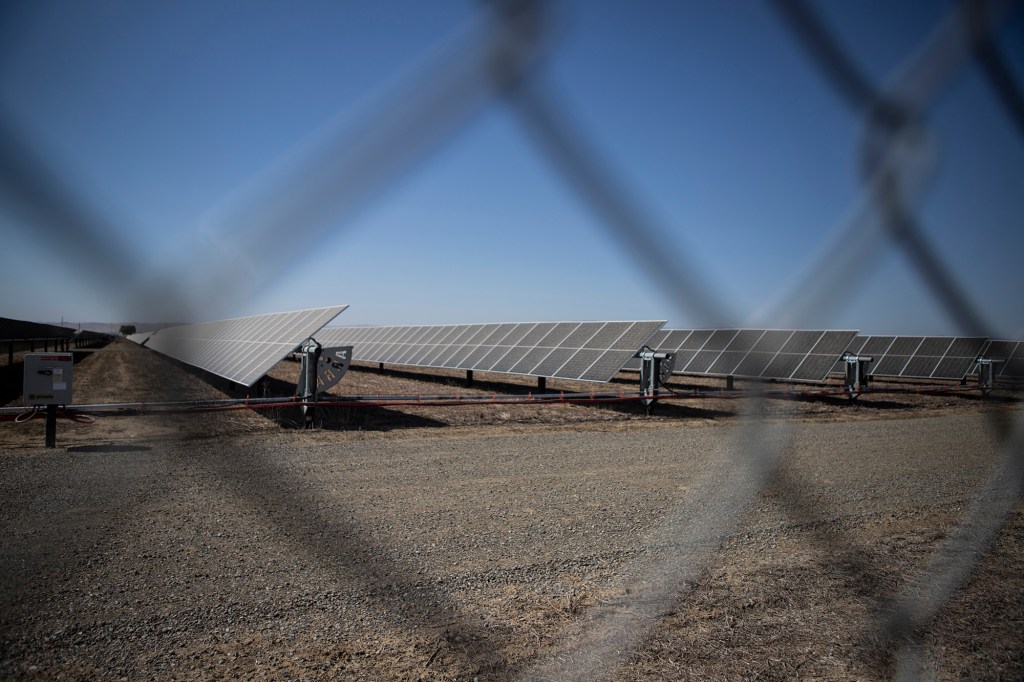 A view of rows of solar panels inside a solar farm is shown through a chain-linked fence on a sunny day.