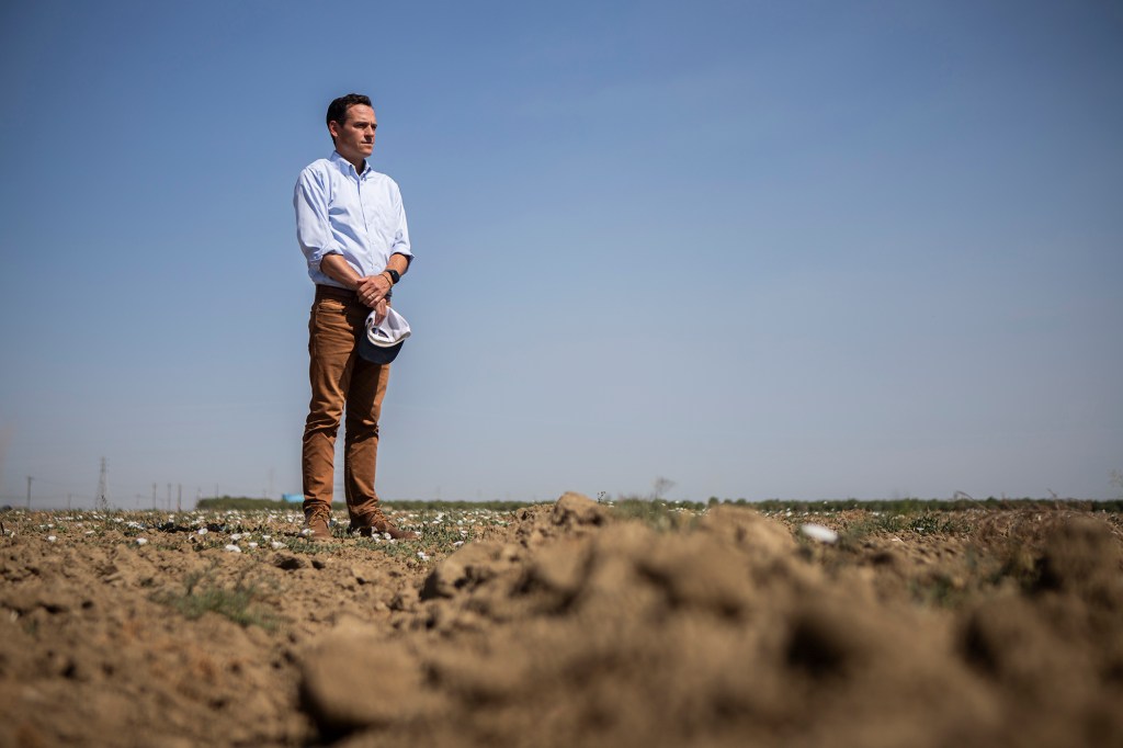 A low-level view of a man in a dress shirt standing in the dirt of a fallow field on a sunny day. The bottom of the frame shows a close-up of the dirt spread out into the field.