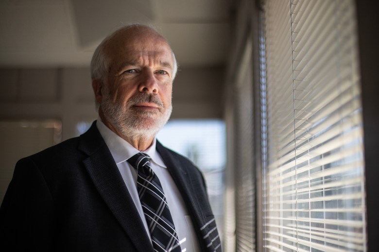 An older person in a suit and striped tie stands indoors beside window blinds, looking ahead with light casting across their face.