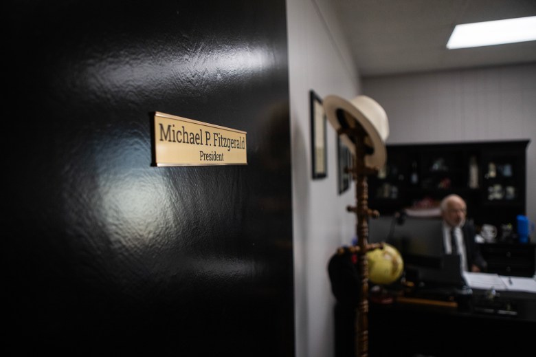 A close-up of a brass nameplate on a dark office door reads “Michael P. Fitzgerald, President,” with the blurred view of a person seated at a desk inside the office in the background.