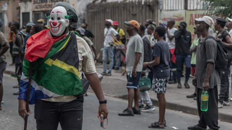 A masked man draped in a South African flag stands in the street as onlookers gather behind him, holding a slingshot in one hand and a stick in the other.r