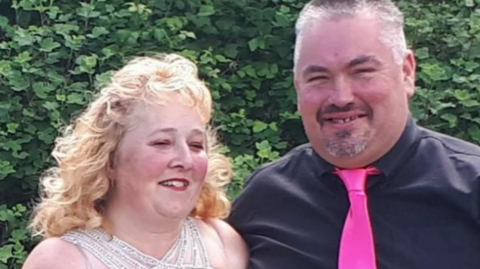 Karen and David Cozens stand next to each other and laugh at an outdoor event. Karen wears a dress and David wears a black shirt and pink tie.