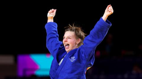 Summer Shaw, in a blue judo robe, lifts her arms in the air after winning Commonwealth Games bronze