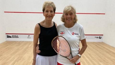 Anne Manley with her arm around a friend on a squash court. She is wearing an England Squash kit and holding a racket