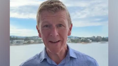 Tim Peake is standing close to the camera, centred in the frame, wearing a light blue button-up shirt. Behind the person is a large body of water with a shoreline and buildings visible in the distance, while the sky is bright with a mix of blue patches and thick white-grey clouds.