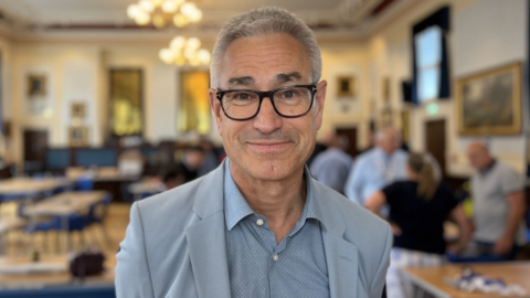 Gerald Voisin stands on a large ornate room. He has short grey hair, wears dark-framed glasses, a light blue blazer, and a pale blue collared shirt.