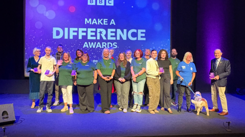 A gathering of people on a stage lit by purple lights and the words Make A Difference Awards on a large screen behind them.