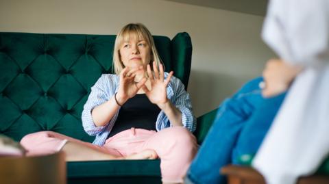 A woman sits in a large dark green, button-tufted armchair during a conversation. She is wearing a light blue and white striped shirt over a dark top and pale pink trousers, with both hands raised as if explaining or demonstrating something. In the foreground, out of focus, another seated person can be seen wearing blue clothing and holding a sheet of paper.