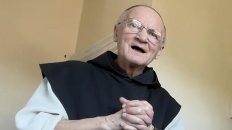 Brother Titus Keet is looking down at the photographer, with his hands clasped. He is wearing monk's robes - a white tunic with a black scapular. He is open-mouthed as if talking. He wears glasses and has very little hair.