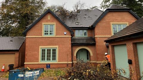 A two-storey new-build detached house in Darlington. It is red-brick and mimics a cottage style, with sections either side of an arched entrance porch. The door, windows and double garage doors are all green. The garage sits at a right angle to the front of the house, obscuring the downstairs window on the right. The garage for presumably a neighbouring property sits to the left of the house and has a brown roller-shutter door. A workman in an orange hi-vis and white hard hat has his back to the camera and appears with a wheelbarrow in the small gap between the house and its double garage. High trees can be seen behind the house, one of which has partially collapsed into the roof, creating a large hole in it. Cut branches are piled up in the front garden