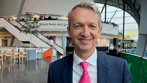 Andy Haldane smiling into the camera. He has grey, short hair and is wearing a blue suit, white shirt and pink tie. He is standing on the concourse of the Glasshouse in Gatesheead which is an open atrium with a white staircase and balcony behind him. There is a cafe with wooden chairs to the left.