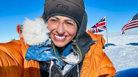 Chandi in a grey wooly hat and a big orange coat in the South Pole. British and USA flags wave on flagpoles in the background.