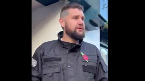 A man with a beard wearing a black jacket with a poppy on it. He is looking away from the camera.