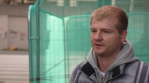 Artem Semchik, wearing a grey hooded top, speaks to the camera in front of green safety netting