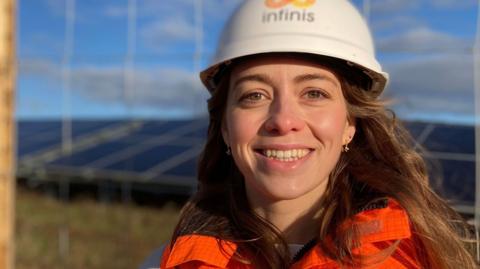 Ellie Goodchild, standing outside, by a solar farm being built behind her. She is smiling and has long hair, is wearing a hard hat and orange high-vis jacket. She is by a fence.