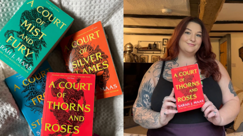 A photo of a woman with red hair smiling and holding a red book with yellow writing on the front cover. She has got tattoos on and a black tank top.Â