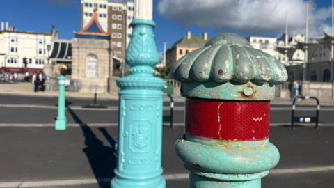 A bollard in the old brighton blue which has faded paint exposing grey cast iron and sections of rust. The original colour is more green than the new paint on a lamp post behind which is freshly painted in a slightly more blue colour near the Palace Pier