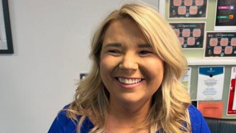 A woman with blonde hair, smiles at the camera. She is wearing a blue dress and you can see the wall of a classroom in the background.