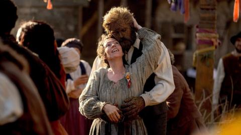 A woman with dark hair and a medieval style dress is embraced by a wicker man.