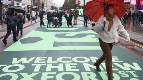 A woman holding red umbrella and others walk along a former roadway on Oxford Street in London.
