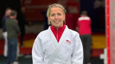 A woman wearing a white judo outfit over a red jacket. She is standing in a room with other people behind her who are out of focus in this shot. She has blonde hair tied in a braid and is smiling for the photograph.
