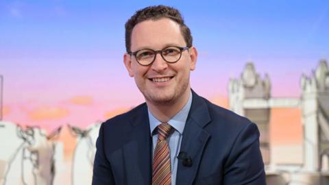 Darren Jones smiles at the camera wearing glasses, a dark blue suit, lighter blue shirt and striped tie. He is wearing a clipped mic on his jacket and he is sitting in a TV studio.
