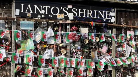 A wall covered in pictures and a string of Welsh flags. At the top a board reads Ianto's Shrine in white writing. The pictures are attached to a rusty metal grid.