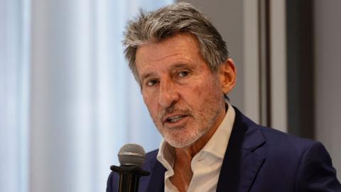 Sebastian Coe, visible from shoulders up wearing a white open-necked shirt and a navy blue suit jacket, holds a microphone up to his mouth as he speaks at a news conference. A wall and a window can be seen out of focus behind him