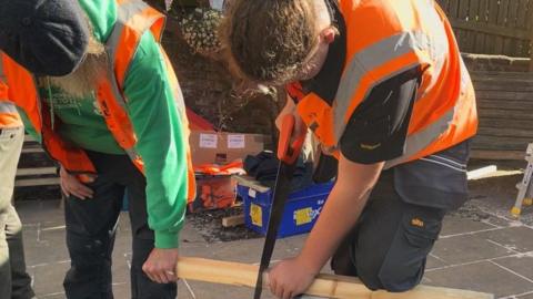 Picture shows two males sawing a piece of light brown timber. Man on the right who has a grey beard is wearing dark trousers, green jumper with a high vis jacket over the jumper and wearing a blue cap is leaning over the wood with his left hand on the timber. The male student is on the right, bent over with a saw in his right hand and is sawing the timber with his left hand and left knee on the wood holding in place.