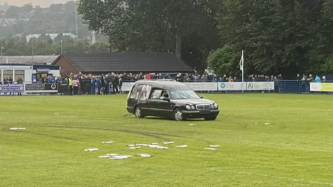 The abandoned hearse in the middle of the grass pitch, covered with tyre tracks, and a scattering of pamphlets on the ground. Football supporters are huddled around the perimeter of the pitch. It was an overcast day.