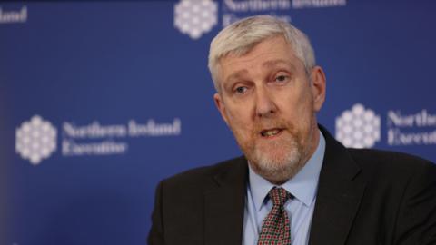 John O'Dowd, a man with short grey hair and stubble, wearing a blue shirt, navy and red tie and black suit jacket. There is a blue wall behind him with the Northern Ireland Executive logo on it. 