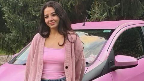 A woman with long, dark hair stands in front of a pink car. She wears a pink top under a pink cardigan and blue jeans.