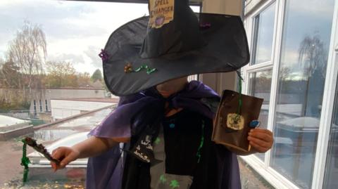 Child dressed as a witch in a homemade Halloween costume featuring a black hat, purple cape, wand and handmade accessories.