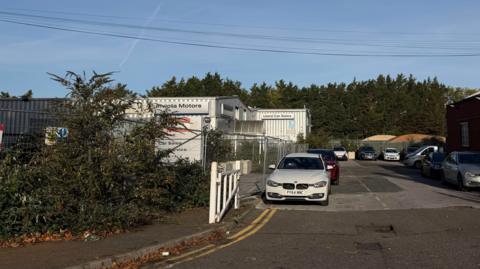 A building brandished with Invicta Motors logos sits on a fenced off site in Sturry Road in Canterbury, Kent. Cars are parked along the road in front of the site underneath a clear sky.