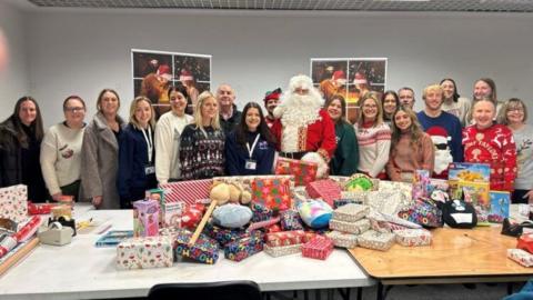 A large group of people are standing in front of large tables that are covered in dozens of wrapped gifts. Standing in the middle of them is Father Christmas, who has a large while curly beard and red and gold outfit.