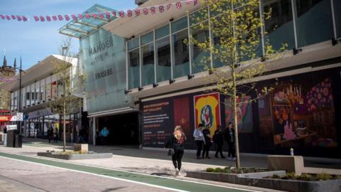 A street with the glass front of the Mander centre on the right with a blue sky behind and Union Jack bunting. The back of a shopper can be seen and a group of several people are walking past a sign with colourful artwork advertising Wolverhampton Art Gallery.