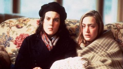 Two people sit closely together on a floral-patterned sofa indoors, wrapped in blankets and warm clothing, facing forward in a softly-lit room.
