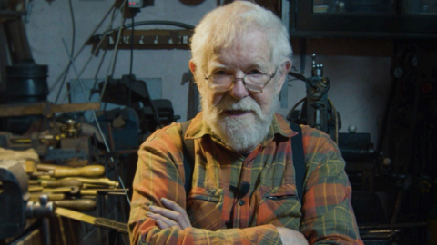 Frank Lee smiling into the camera with his arms crossed in front of his chest. He has short, white hair and a white beard and is wearing rectangular glasses. He is wearing an orange and green checked shirt with black braces around his shoulders. He has a small black microphone clipped to his chest. He is standing in his workshop and behind him there is an array of tools and equipment with wooden handles. It is very cluttered.
