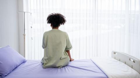 A woman sits on a hospital bed. She has her back to the camera. The room is featureless, white and sterile.
