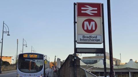 A public transport terminus. A large red and white sign indicates it's Bradford Interchange. The sign has a train track symbol on it, and a large white 'M' in a red circle for Metro. To the left of the picture, a bus is parked up. To the right, slightly in the background, is a concourse with canopies and a large sign at the entrance also saying Bradford Interchange.