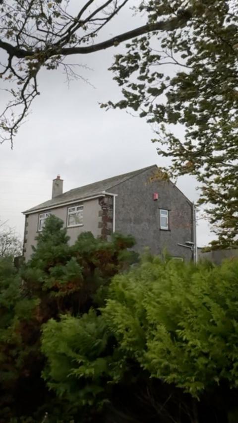 A two-storey grey house with a pitched roof sits partially hidden behind dense green shrubs and trees. Overhanging branches frame the top of the image, and the house is viewed from a slightly lower angle against a cloudy, overcast sky. The scene appears rural and surrounded by mature vegetation.
